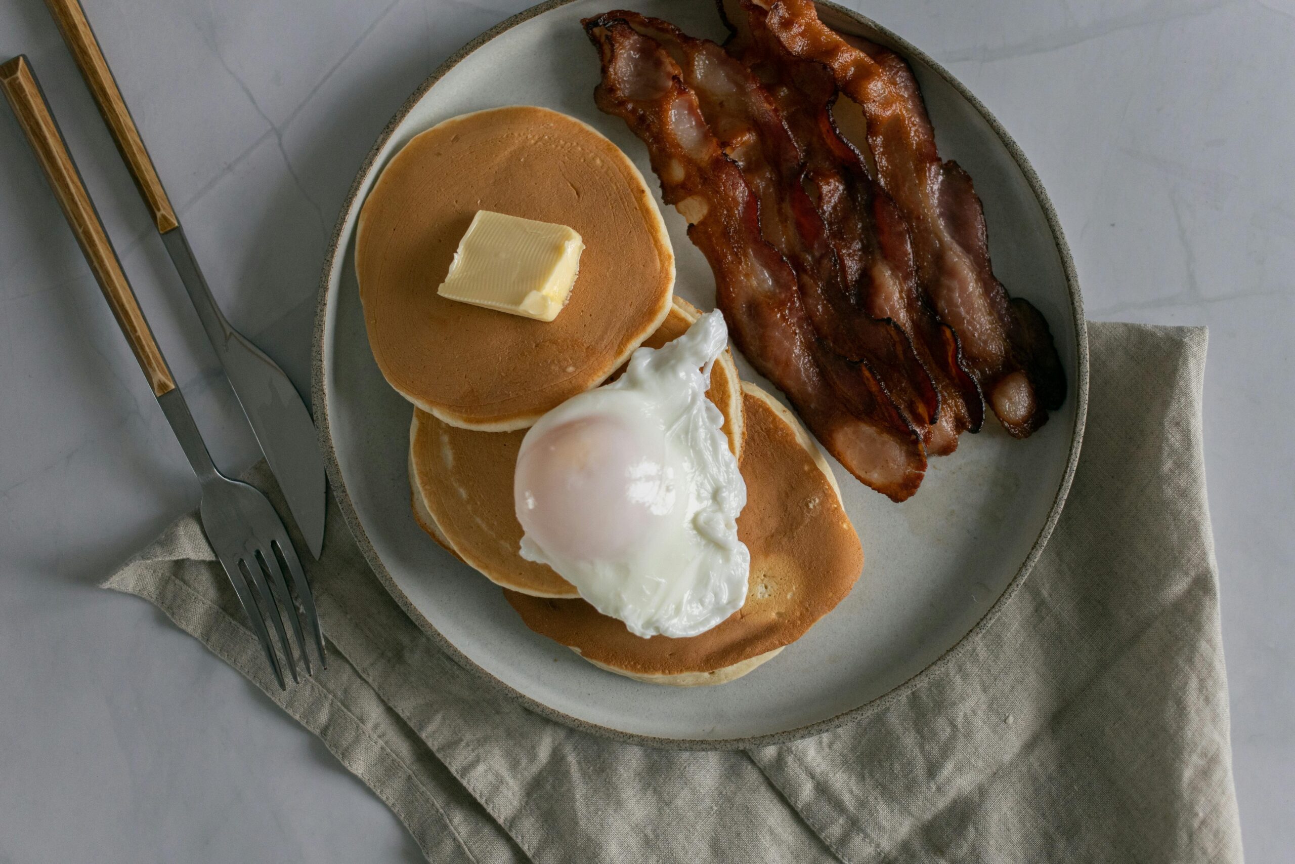 Top view of delicious pancakes with butter piece and poached egg near sliced fried bacon on plate near fork and knife near napkin on white table in bright place
